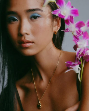 Close-up portrait of a woman with blue eyeshadow and vibrant magenta orchids held near her face, featuring gold jewelry.