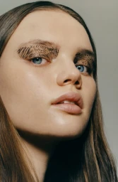 Close-up portrait of a young woman with intricate graphic eye makeup and blue eyes against a plain grey background