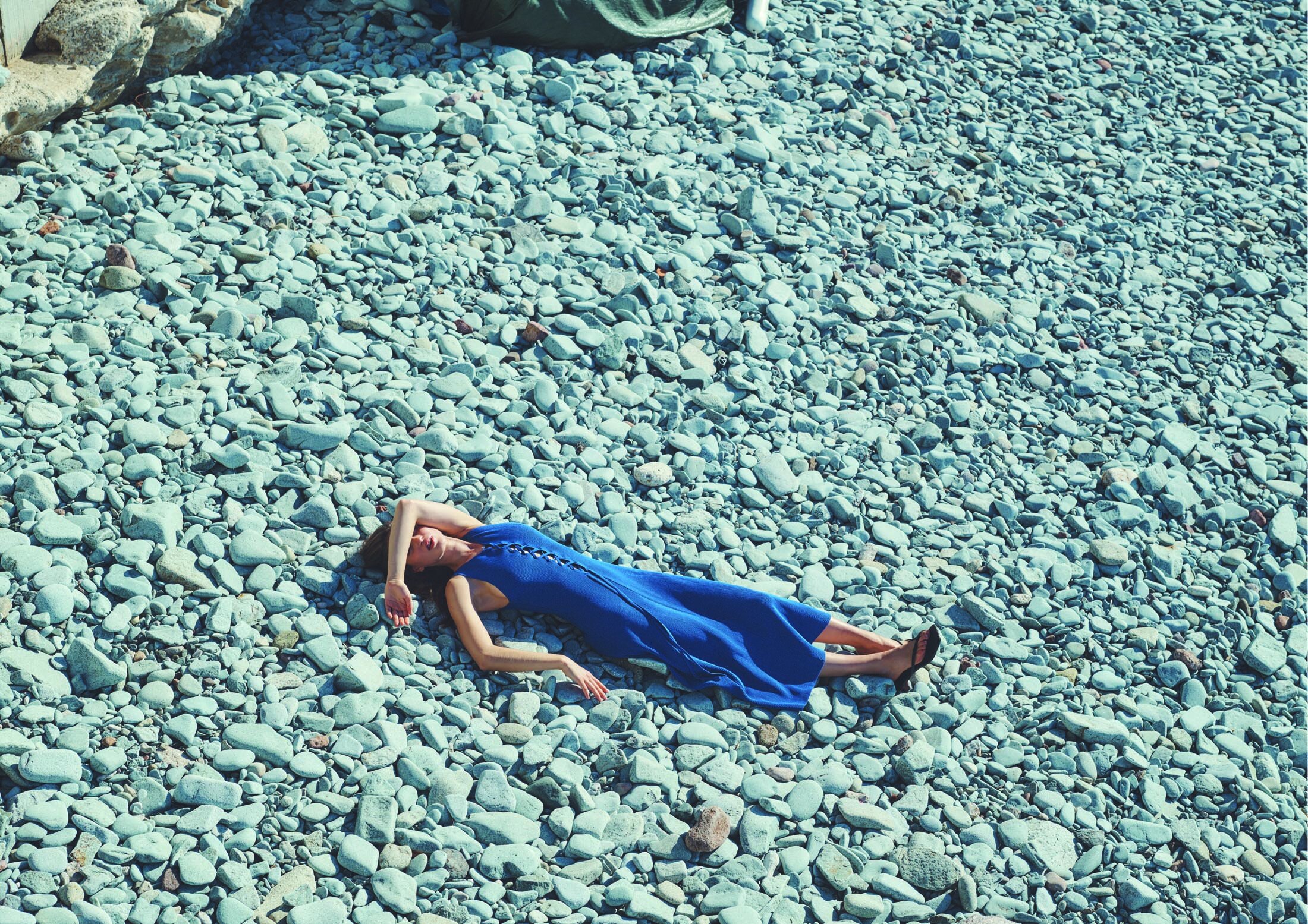 Woman in blue dress lies on light grey pebble beach with arm over face, enjoying sun. Natural light, fashion and relaxation.