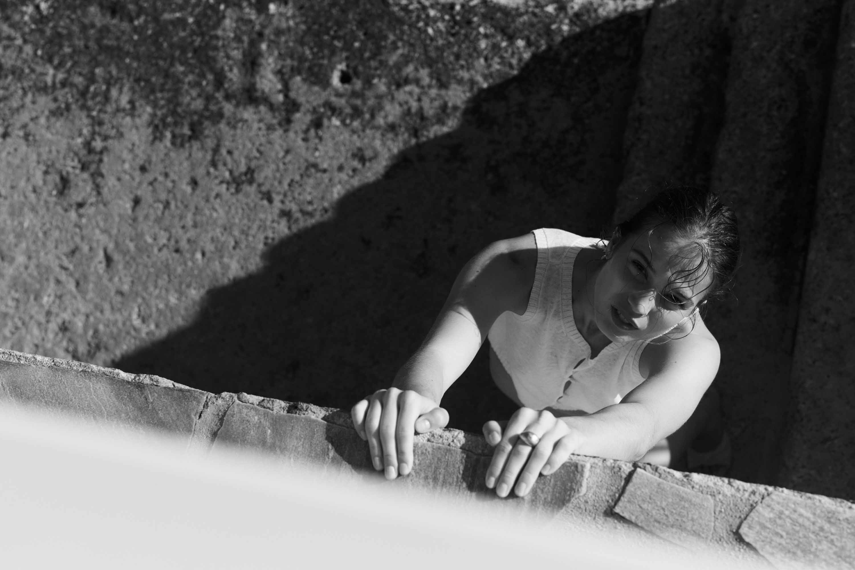 Monochrome portrait of a woman leaning over a stone wall, captured from a high angle with natural lighting and textured background.