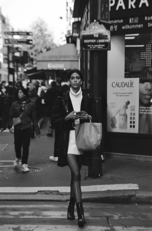 Black and white street photograph of a woman walking across a busy city street holding a shopping bag and smartphone.