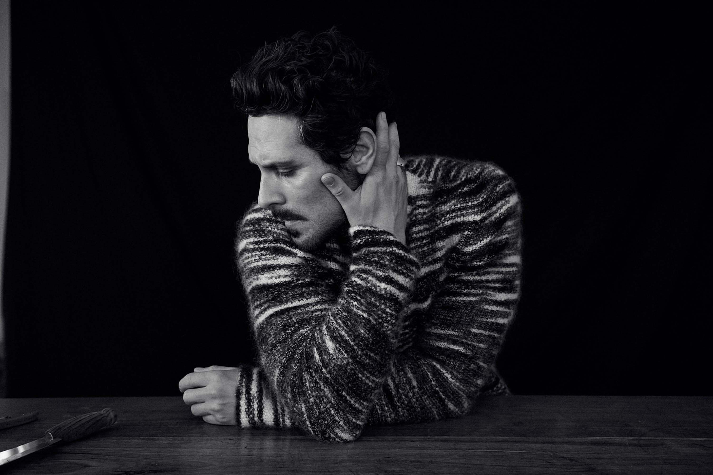 Black and white portrait of a man with dark hair and a mustache wearing a patterned sweater, resting his hand against his face.