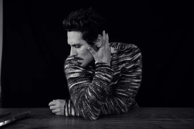 Black and white portrait of a man with dark hair and a mustache wearing a patterned sweater, resting his hand against his face.