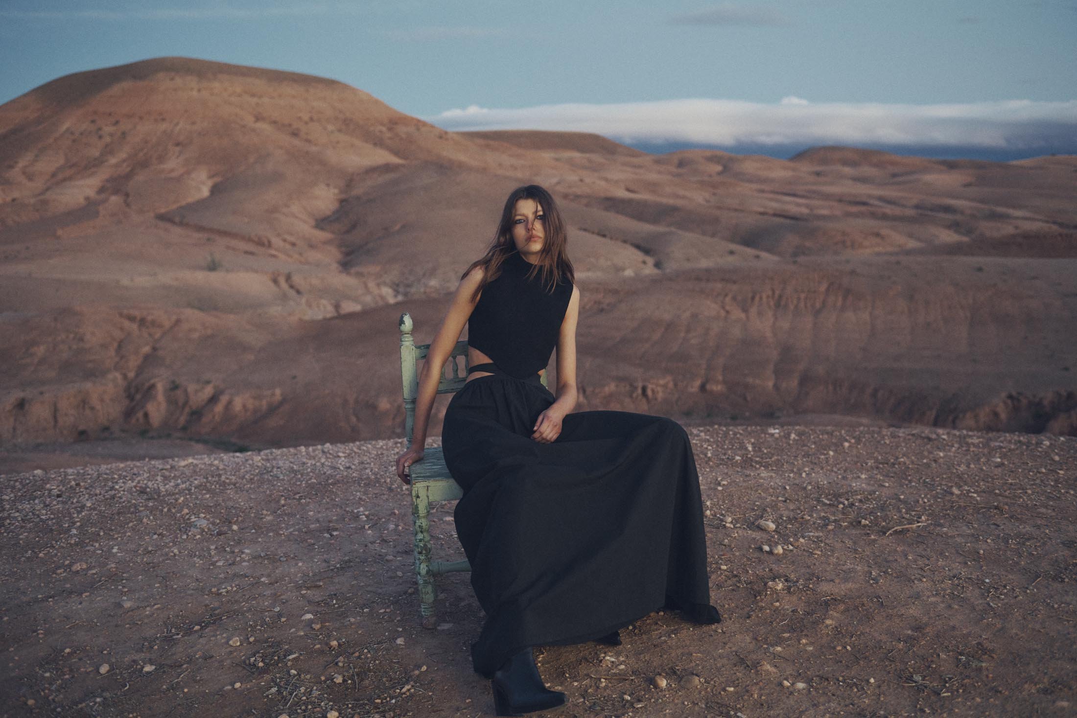 A brunette woman in a black outfit sitting on a green chair with a desert landscape in the background underneath a clear sky.