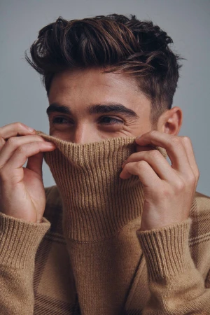 Close-up portrait of a young adult male with brown hair partially covering his face with a tan turtleneck sweater against a grey backdrop.