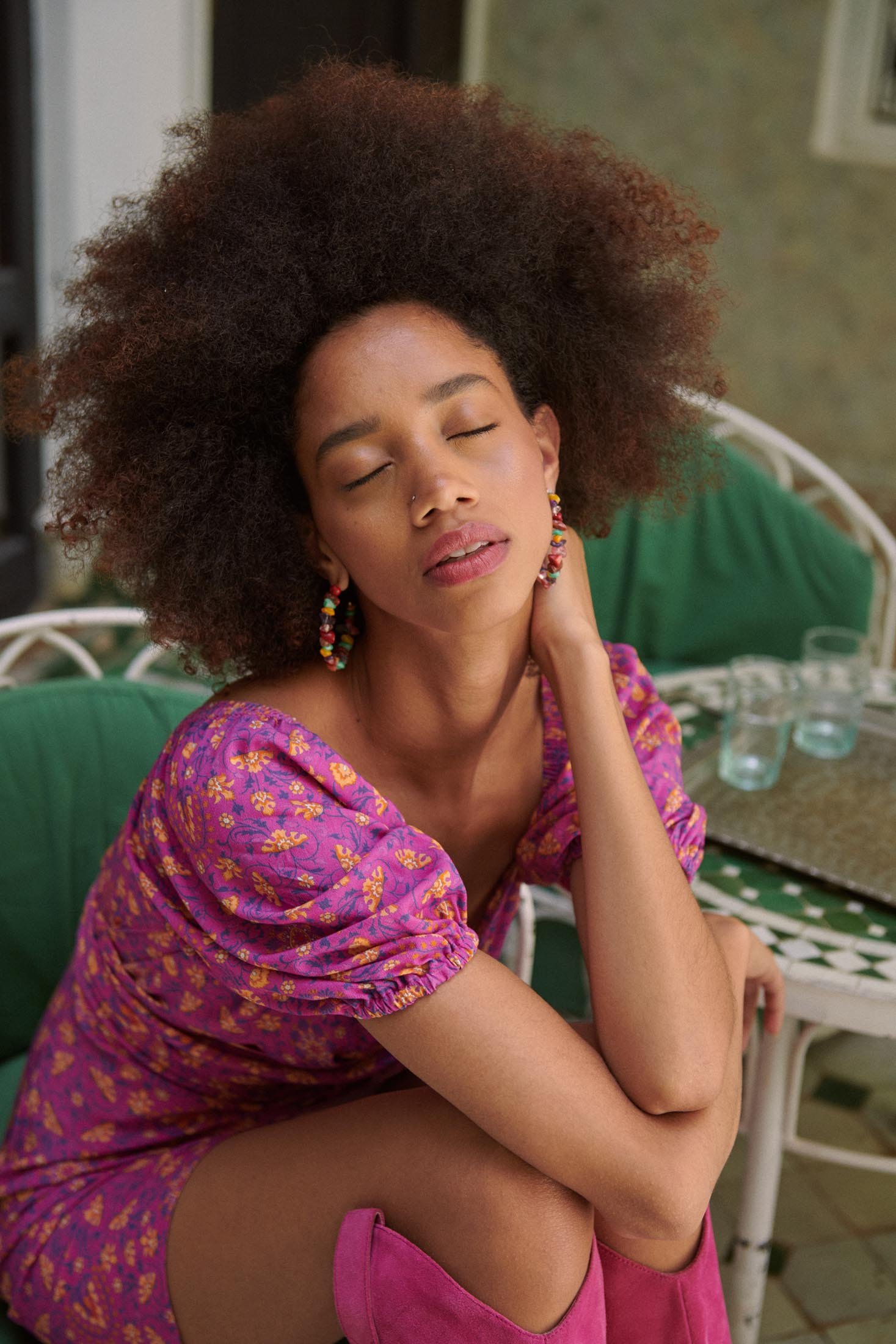 A woman with dark curly hair and wearing a floral mini dress poses indoors with her eyes closed and touches her neck gracefully.