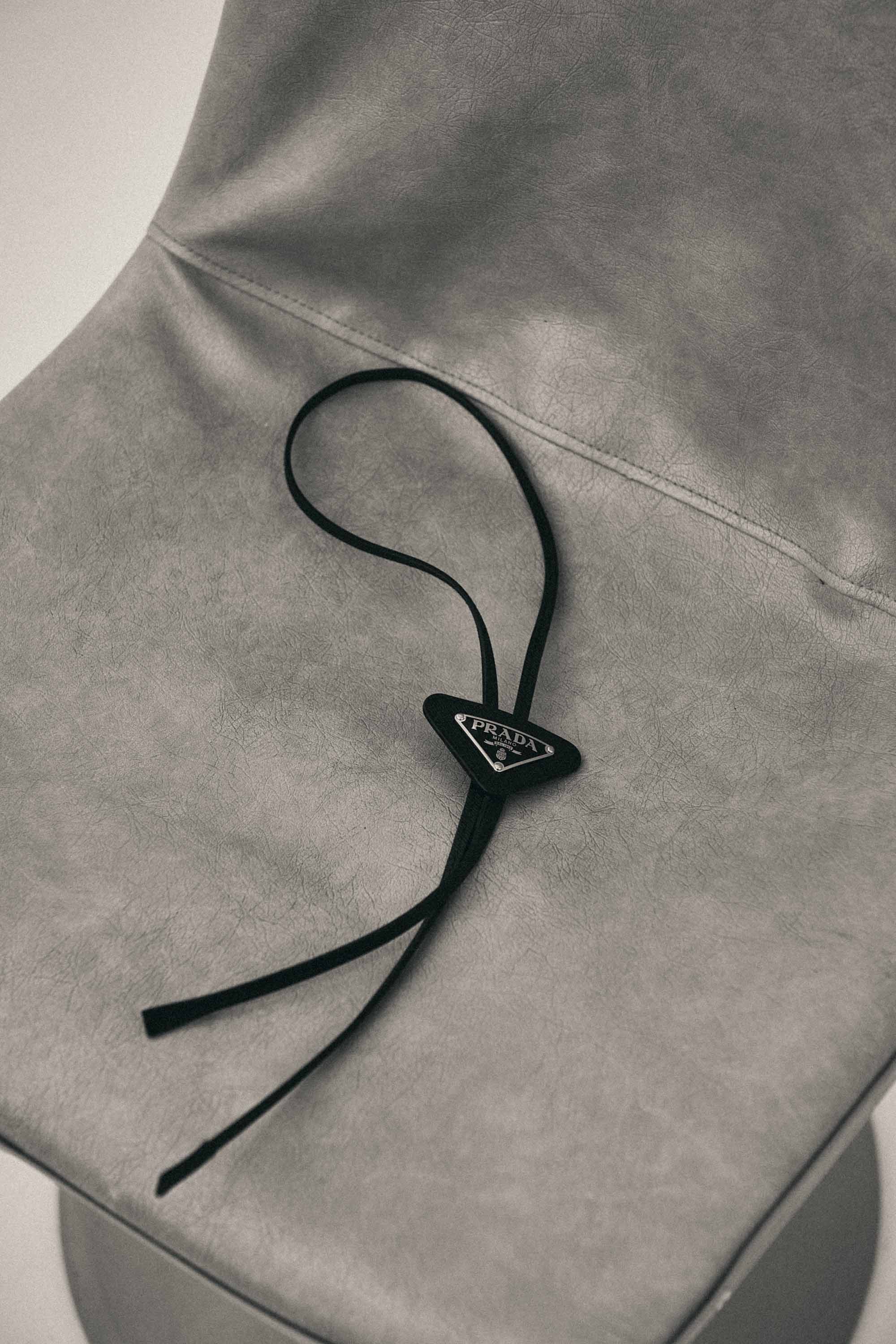 Close-up of a grey upholstered chair with a black Prada bolo tie resting on the seat. Minimal composition, simple background.