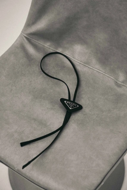 Close-up of a grey upholstered chair with a black Prada bolo tie resting on the seat. Minimal composition, simple background.