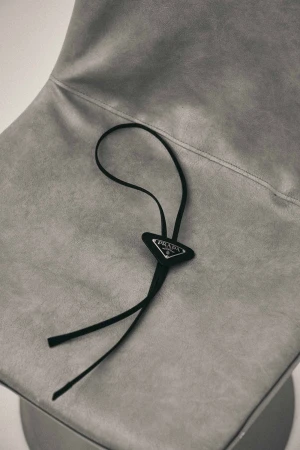 Close-up of a grey upholstered chair with a black Prada bolo tie resting on the seat. Minimal composition, simple background.