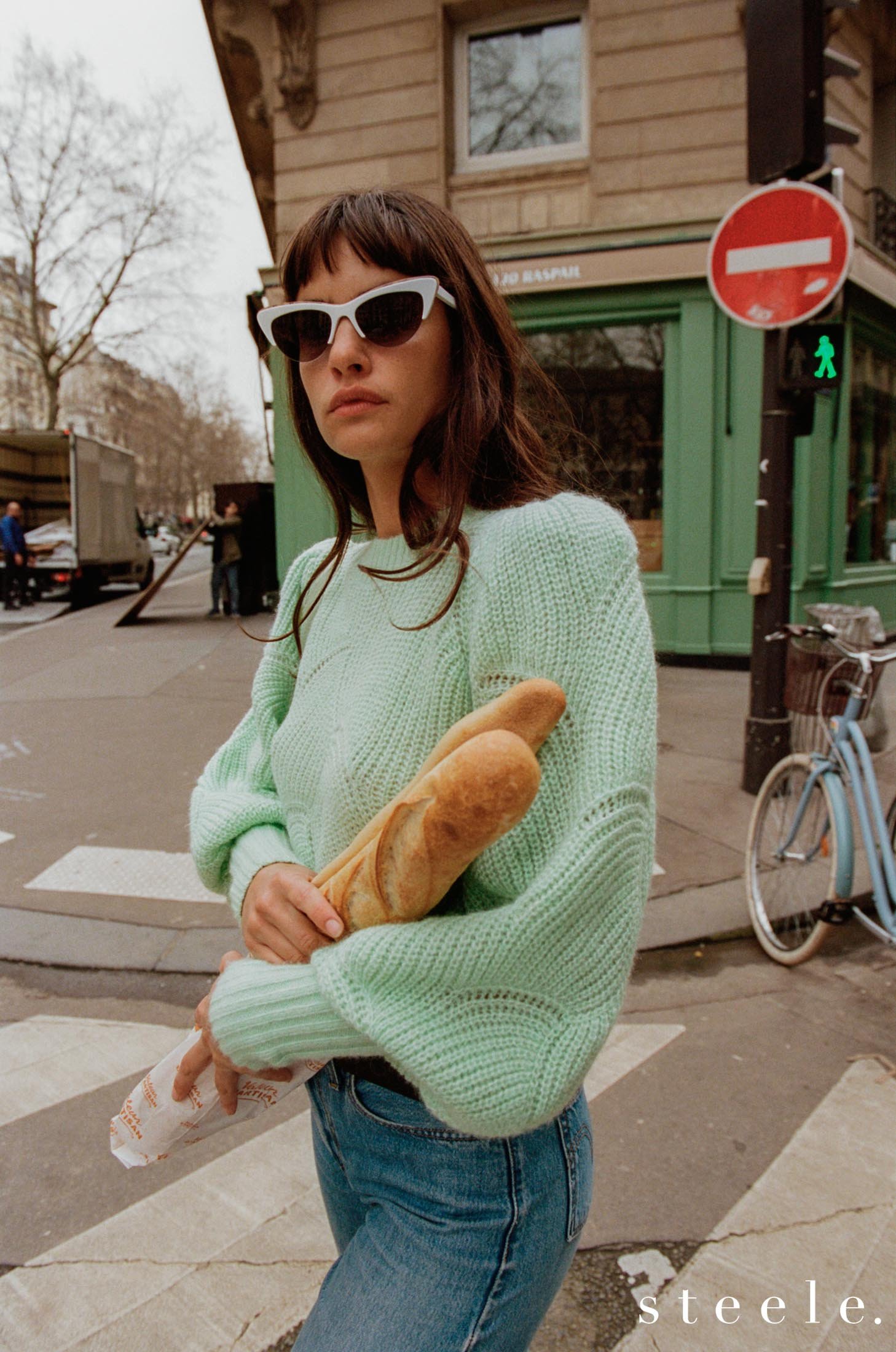 A woman stands on a crosswalk in an urban setting, wearing sunglasses and a green sweater, holding baguettes. The scene exudes casual, fashionable style.