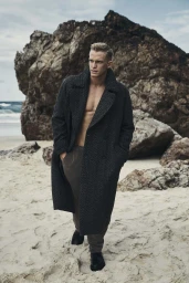 Blonde man wearing a dark textured oversized wool coat and trousers standing on a sandy beach