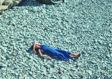 A woman in a vibrant blue dress lying down on a bed of smooth grey river stones and rocks.