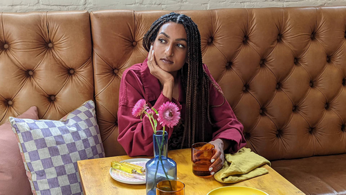 Young woman with braided hair sits at a table with flowers in a vase and a drink in an amber glass in an indoor restaurant setting.