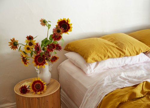Still life of sunflowers in a white vase on a round rattan table, set against a white wall near a bed with yellow and white pillows.