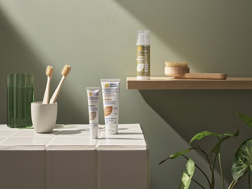 Still life of skincare products, toothbrushes, glass of water, and plant against a green wall in a bathroom setting, studio lighting.