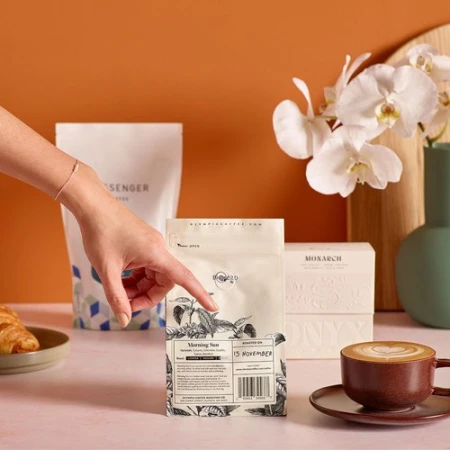 Close-up of a hand pointing to a bag of Morning Sun coffee with latte, croissant, and orchids, on a peach surface and orange backdrop.