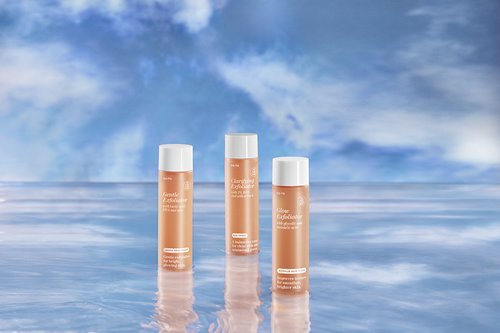 Three bottles of skincare product stand in front of a blurred blue and white sky and water background in a beauty product shot.