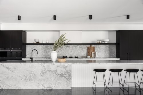 Modern kitchen interior featuring a marble island with black stools, black cabinets, white backsplash and black lighting fixtures.