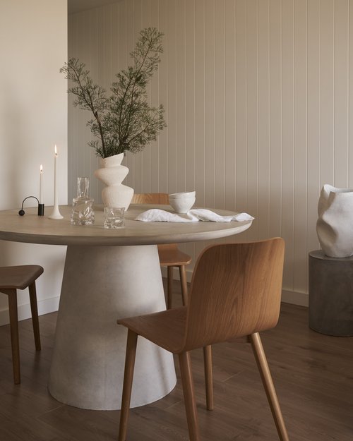 Interior view showcases a round dining table with a decorative vase, clear glassware, and wooden chairs against a light backdrop.
