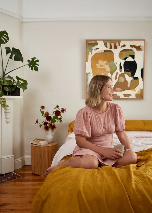 Blonde woman in pink dress sits on mustard bed holding mug in white bedroom with abstract art and monstera plant.