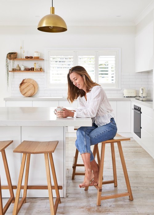 Woman sitting at a white kitchen island with a cup, illuminated by a gold pendant light and natural light from a window.