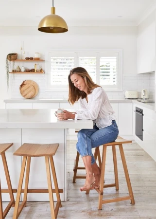 Woman sitting at a white kitchen island with a cup, illuminated by a gold pendant light and natural light from a window.