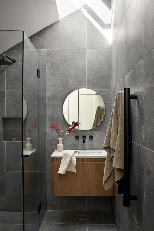 Modern bathroom interior with grey tiles, wooden vanity, circular mirror, and black fixtures under a bright skylight.
