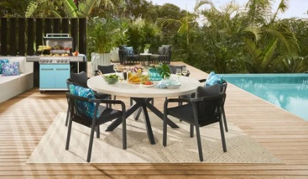Outdoor dining set on a wooden deck by a swimming pool, styled with a round table and black armchairs.
