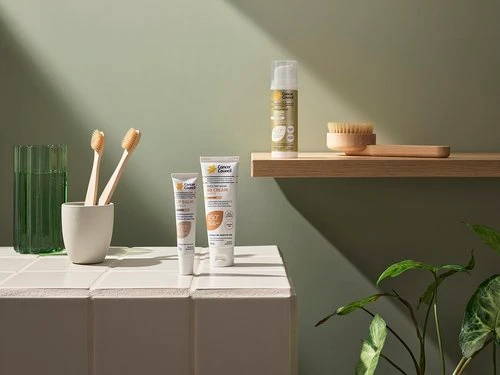 Skincare product tubes, wooden toothbrushes in a holder, and a floating shelf styled against an olive green wall.