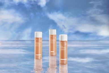 Three orange cylindrical skincare bottles standing upright on a reflective water surface against a blue sky backdrop.