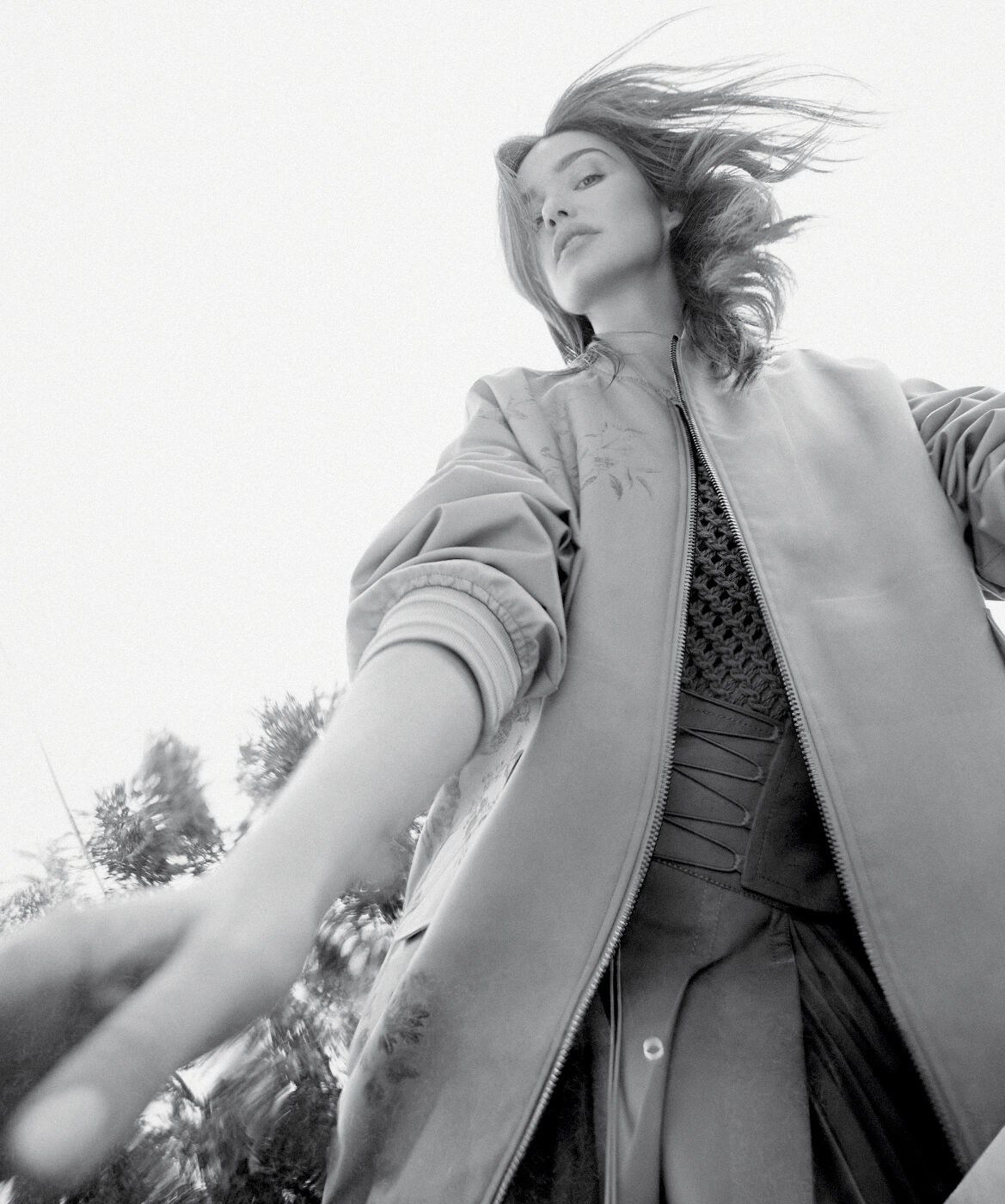 Monochrome low-angle shot of a woman in a jacket with wind-blown hair, looking down with a blurred arm reaching toward the camera.