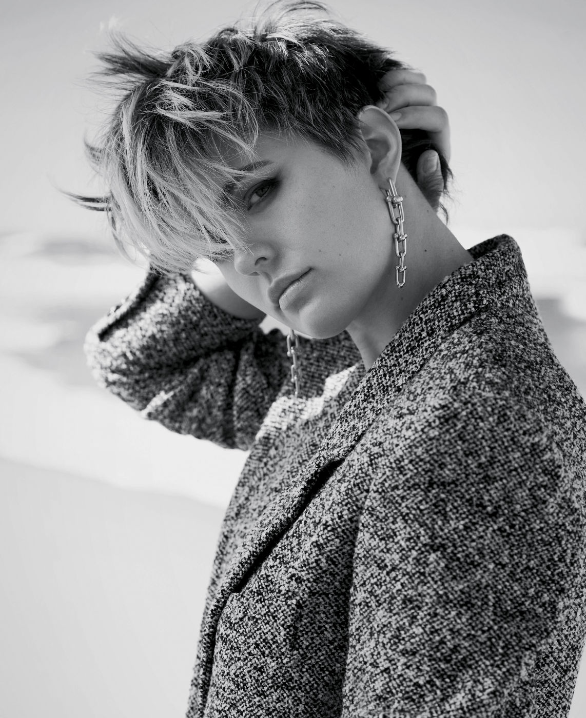 Monochrome portrait of a stylish woman with short hair and an oversized textured grey coat, accessorized with statement earrings, posing indoors.