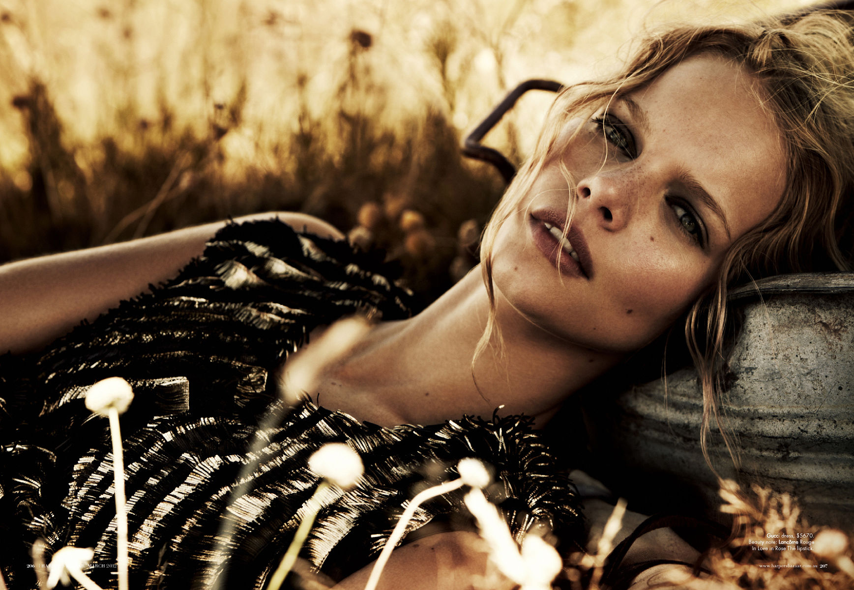 A fair-skinned woman with blonde hair rests her head against a weathered metal container in a sunlit field with delicate white wildflowers.