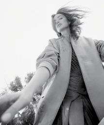 Low-angle monochrome portrait of a woman wearing a bomber jacket outdoors with sky and treetops visible behind her
