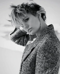 Black and white portrait of a person with short tousled hair wearing a textured tweed jacket and drop chain earrings.