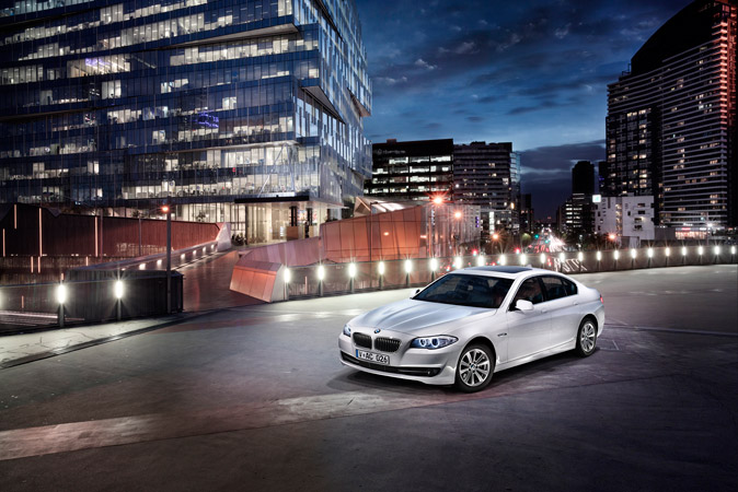 A white BMW sedan is parked on a concrete surface in front of a modern building with glass windows at night with city lights.