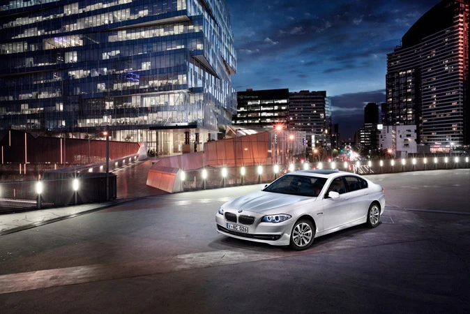 A white BMW sedan is parked on a concrete surface in front of a modern building with glass windows at night with city lights.