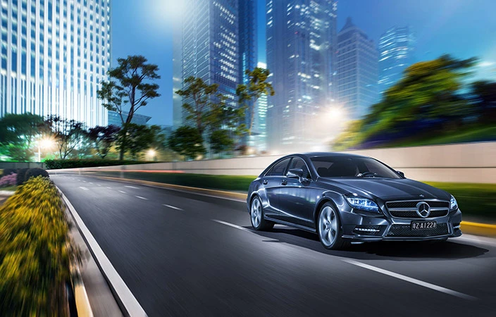 Sleek grey Mercedes-Benz sedan speeds down an urban road at dusk with modern buildings and blurred motion adding a dynamic effect.