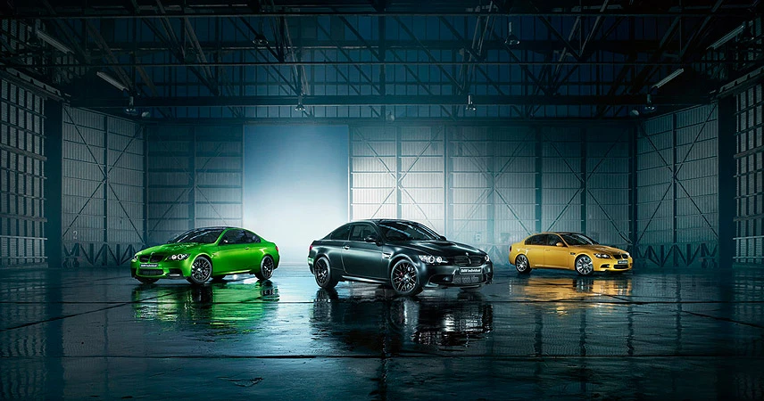 Three BMW M3 GTS cars, in yellow, grey, and green, are parked side-by-side inside a large indoor garage with glossy concrete floors.