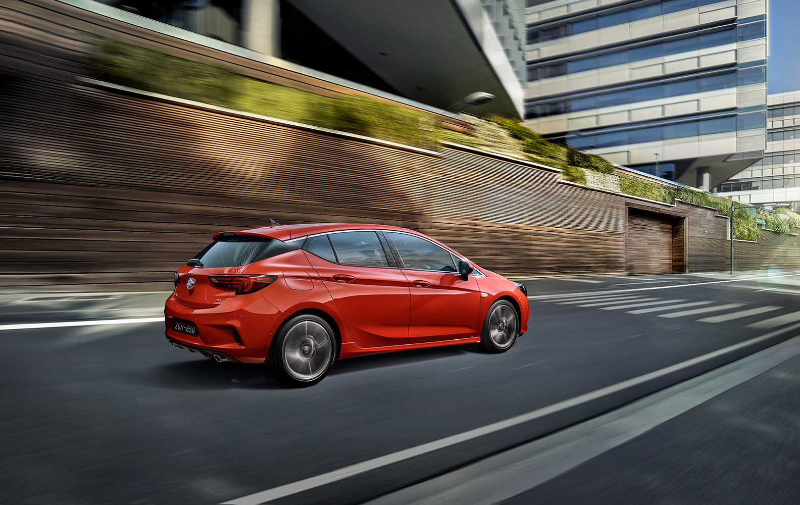 A red hatchback car drives on a city street with blurred motion. The modern architecture includes a wall with green foliage.