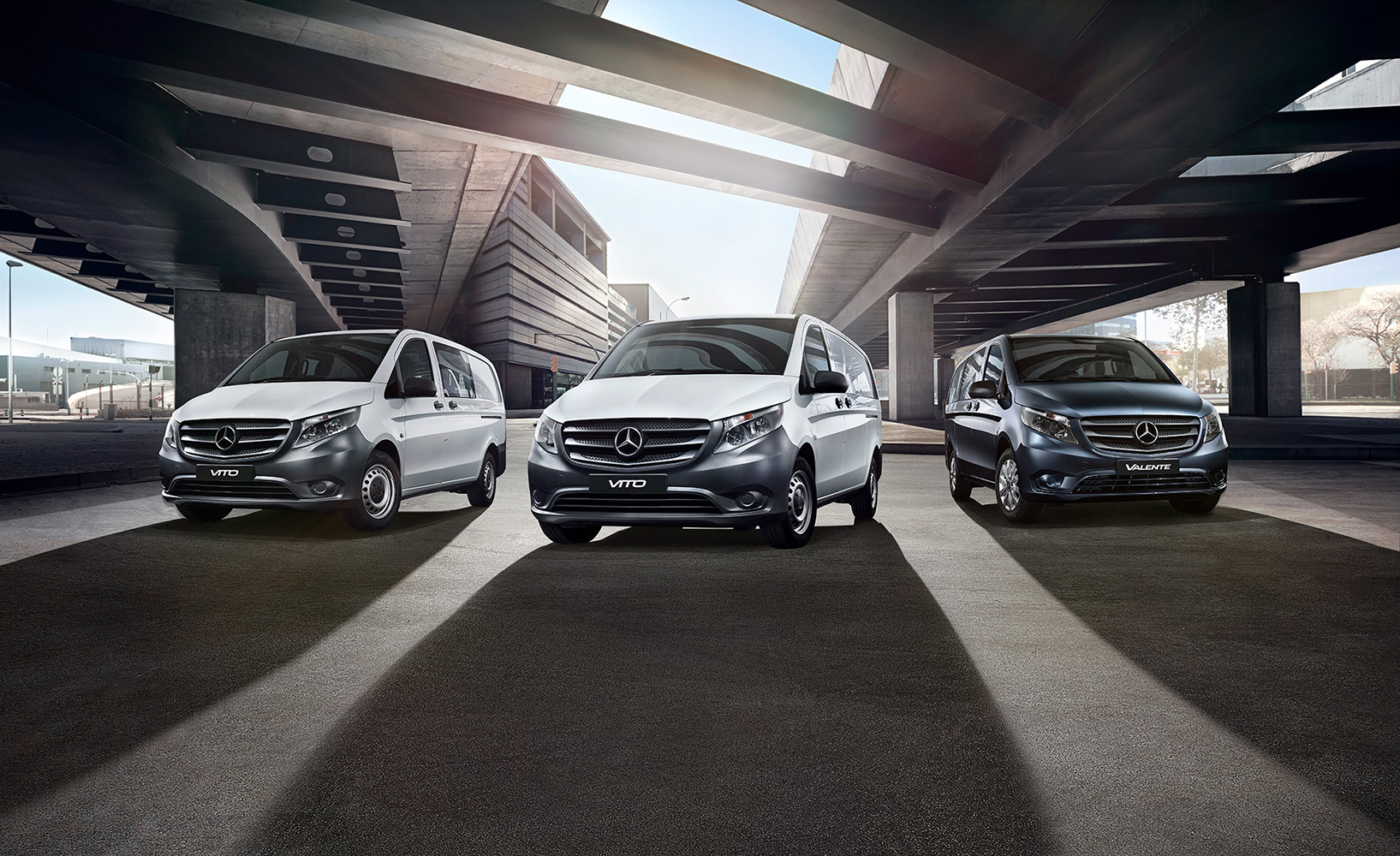 Three Mercedes-Benz vans, in white and dark grey, parked under a concrete structure. Automotive photography with modern architecture in the background.
