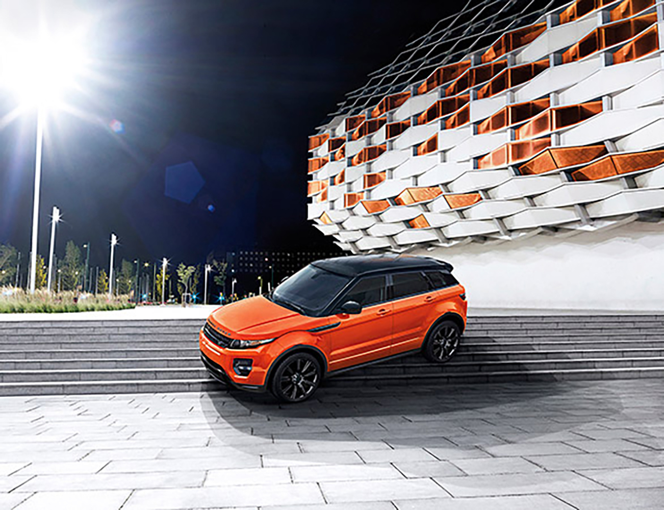 Orange Range Rover SUV parked on stone steps near a modern building with patterned facade, under a bright spotlight at night.