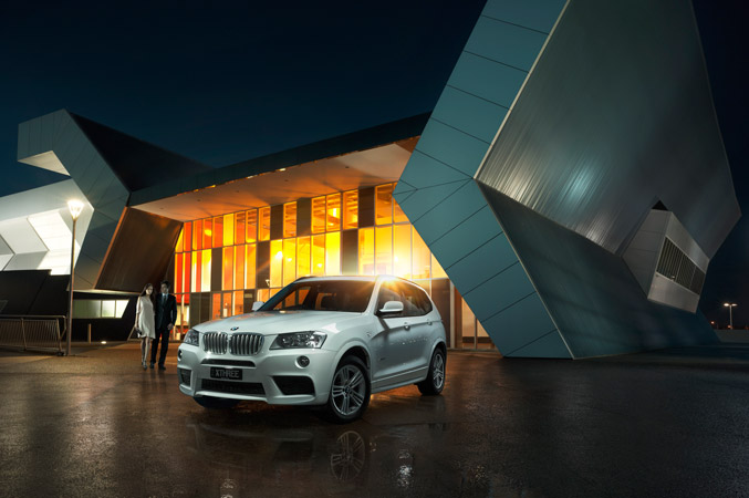 White BMW X3 parked in front of modern building at night with a couple. Automotive luxury car photography for Visie creative platform.