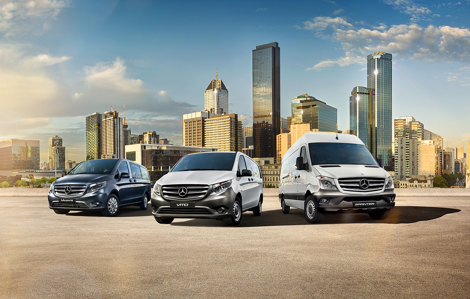Three Mercedes-Benz vans are parked in front of a cityscape with various buildings under a partly cloudy, daytime sky.