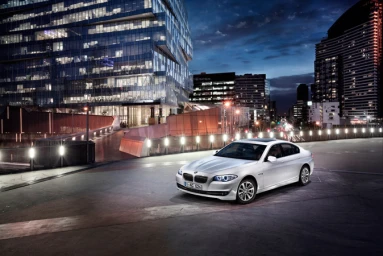 White sedan parked on an asphalt rooftop with modern illuminated glass skyscrapers in the background at night.