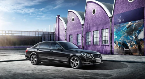 A black sedan parked on a paved ground in front of a purple brick building with distinctive saw-tooth rooflines.