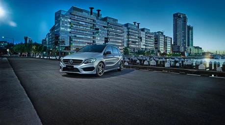 Silver Mercedes hatchback parked on a paved waterfront quay at dusk with modern high-rise buildings in the background.