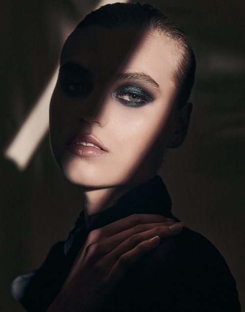 Close-up portrait of a woman with dramatic makeup and a light stripe accentuating her features in a dark, neutral-toned atmosphere.