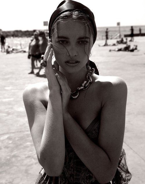 Monochrome portrait of a fair-skinned woman in headscarf and necklace at a beach with other people in the background, gazing at camera.