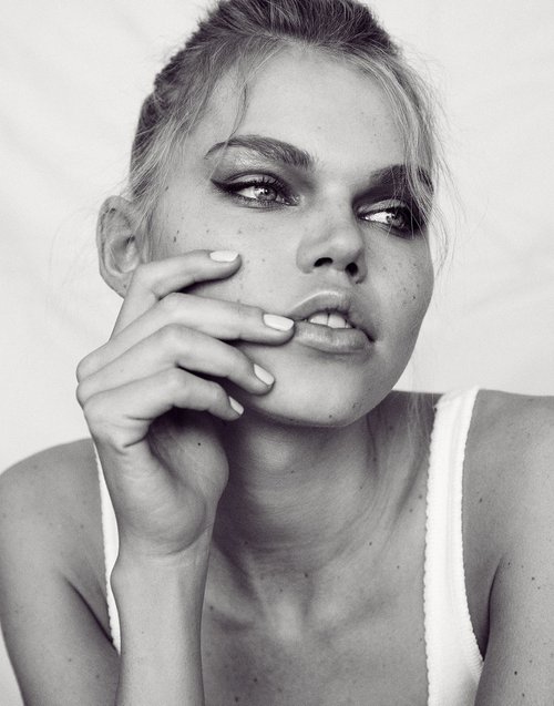 Close-up black and white portrait of a young woman with smokey eyes, her hand near her face, against a bright neutral background.